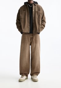 Bruine denimjas met twee voorzakken, gedragen over een zwarte trui en een paar brede bruine jeans. Lichtgekleurde sneakers maken de outfit compleet.