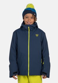 Giacca impermeabile blu navy con una zip e un logo giallo brillante, dotata di cappuccio e polsini elasticizzati, indossata sopra pantaloni gialli.