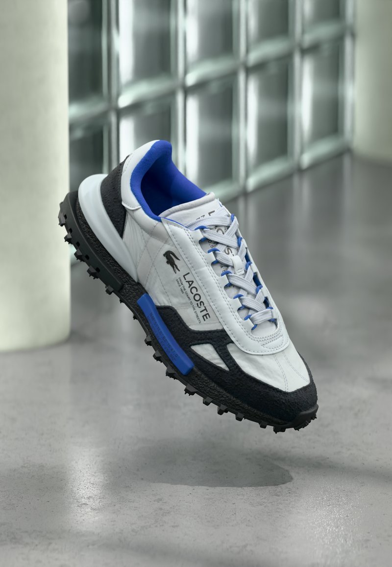 Bela in črna superga Lacoste z modrim notranjostjo in vezalkami, ki lebdi nad betonsko tla blizu stene iz steklenih blokov.