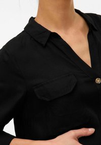 Camisa negra con collar, cierre de botones y un solo bolsillo en el pecho. Tela suave con una textura lisa, que presenta un leve brillo.