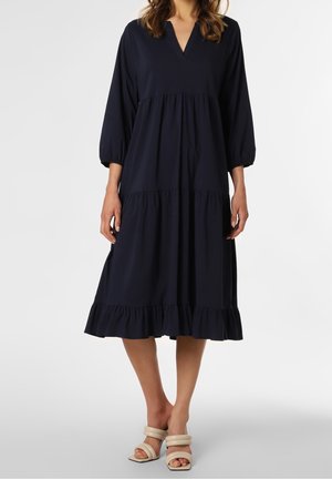 Femme portant une robe midi bleu marine à volants, manches trois-quarts, et des sandales à talons beiges, debout devant un fond uni.