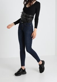 Kvinna som bär mörka tajta jeans, svart långärmad topp, svarta sneakers och en svart strukturerad axelväska mot en enkel bakgrund.