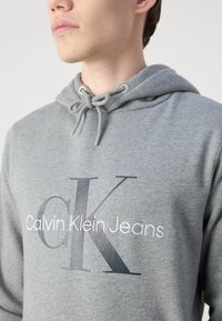 Felpa grigia con cappuccio caratterizzata da un cordino, logo ricamato "Calvin Klein Jeans" in bianco e grigio scuro, realizzata in morbido materiale di cotone con una texture liscia.