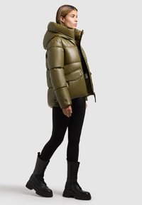 Olivgrüne gepolsterte Daunenjacke mit hohem Kragen, Reißverschluss und zwei seitlichen Taschen. Getragen mit enganliegenden schwarzen Hosen und klobigen schwarzen Stiefeln.