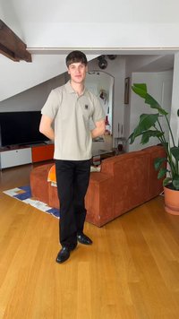 Jeune homme debout dans un salon, portant un polo beige et un pantalon noir, avec un canapé couleur rouille et une grande plante en pot à proximité.