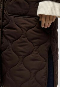 Main reposant sur une veste matelassée brun foncé avec des coutures arrondies, montrant le poignet en tricot blanc et un jean bleu en dessous.