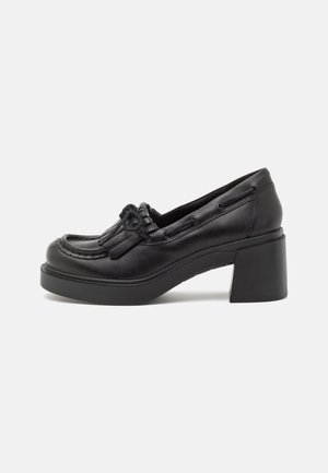 Mocassin pour femme en cuir noir avec talon bloc épais, détail pompon et semelle plateforme sur fond uni clair.