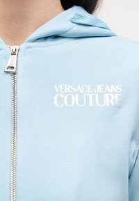 Svetlomodrá mikina na zips s nápisom "Versace Jeans Couture" v bielej farbe na ľavej strane hrude a so zlatým ťaháčikom na zips.