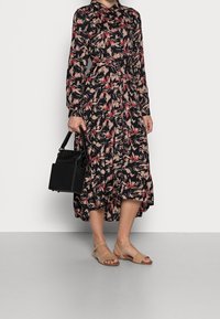 Czarna sukienka z długim rękawem w kwiatowy wzór w odcieniach różu i beżu, z talią podkreśloną paskiem, zestawiona z czarną torebką i beżowymi sandałami.