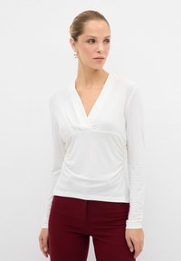 Maglietta bianca a maniche lunghe con scollo a V drappeggiato, caratterizzata da dettagli laterali arricciati. Abbinata a pantaloni aderenti bordeaux. Tessuto dalla consistenza liscia.