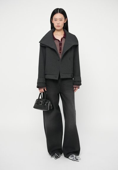 Ženska nosi temno preveliko jakno, široke črne hlače, srebrne špičaste čevlje, drži majhno črno torbico in stoji ob belem ozadju.