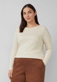 Licht beige lang mouwtop met een ronde hals en ribbeltextuur, gecombineerd met bruin suède-achtige broeken met zijzakken.