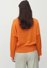 Maglione arancione oversize con polsini e orlo a coste, abbinato a pantaloni grigi chiari con tasche a welt. Tessuto liscio e morbido.