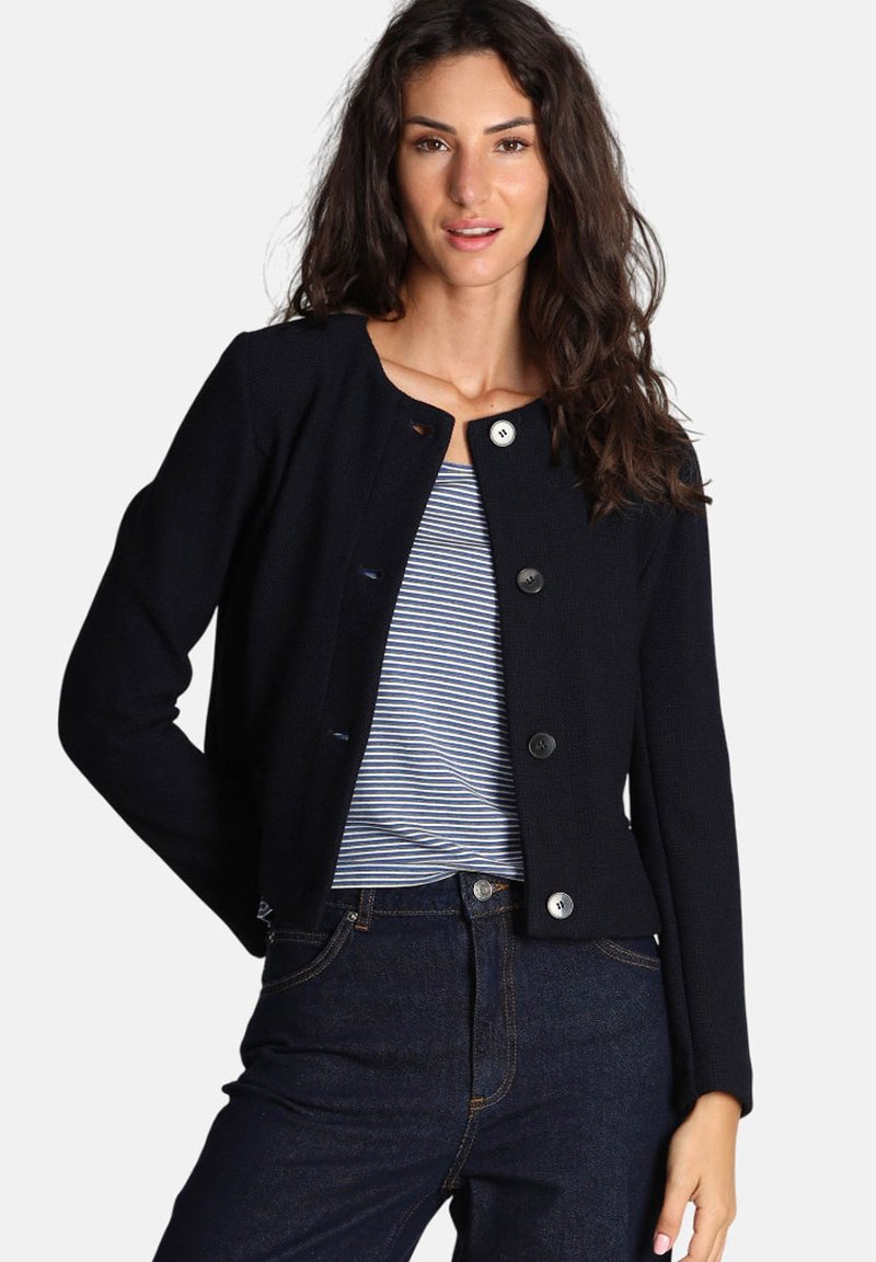 Cardigan lavorato a maglia blu navy con colletto rotondo, chiusure con bottoni, maniche lunghe e design minimalista, abbinato a una maglietta a righe e jeans scuri.