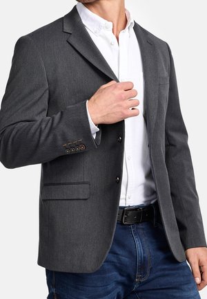 Man adjusting charcoal gray blazer over white button-up shirt, wearing blue jeans and black belt, against white background.