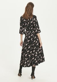 Svart klänning med rosa paisleymönster, tre fjärdedels ärmar och insydd midja, flytande till midikjollängd. Bärs med ankellånga klackar.