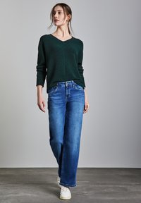 Maglione verde scuro, design a V, tessuto strutturato. Jeans a gamba larga blu con vestibilità classica. Il modello indossa sneaker bianche. Sfondo semplice.