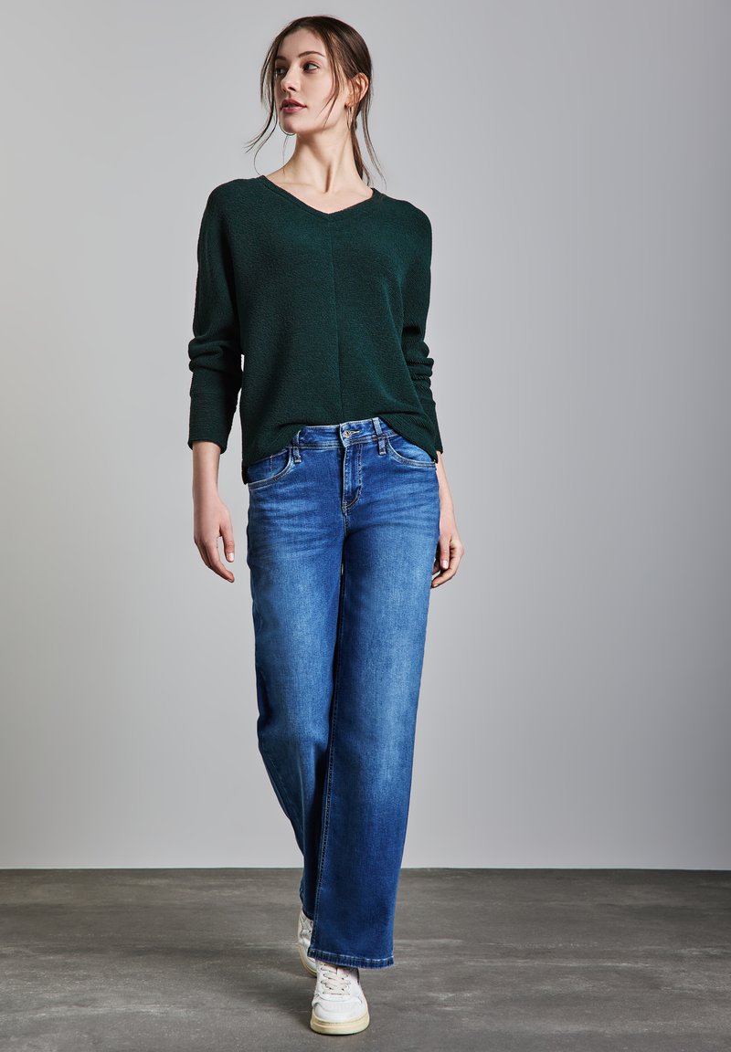 Maglione verde scuro, design a V, tessuto strutturato. Jeans a gamba larga blu con vestibilità classica. Il modello indossa sneaker bianche. Sfondo semplice.