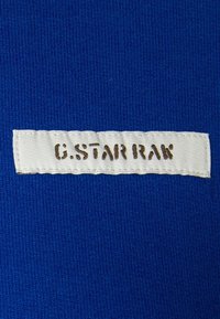 Modrá látka se strukturovaným povrchem. Obsahuje bílou cedulku přišitou s nápisem "G.STAR RAK" v hnědých písmech. Cedulka je umístěna uprostřed.