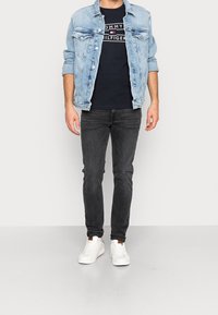 Lyseblå denimjakke over en navy Tommy Hilfiger t-shirt, parret med mørke jeans og hvide sneakers. Casual fit, minimalistisk design.