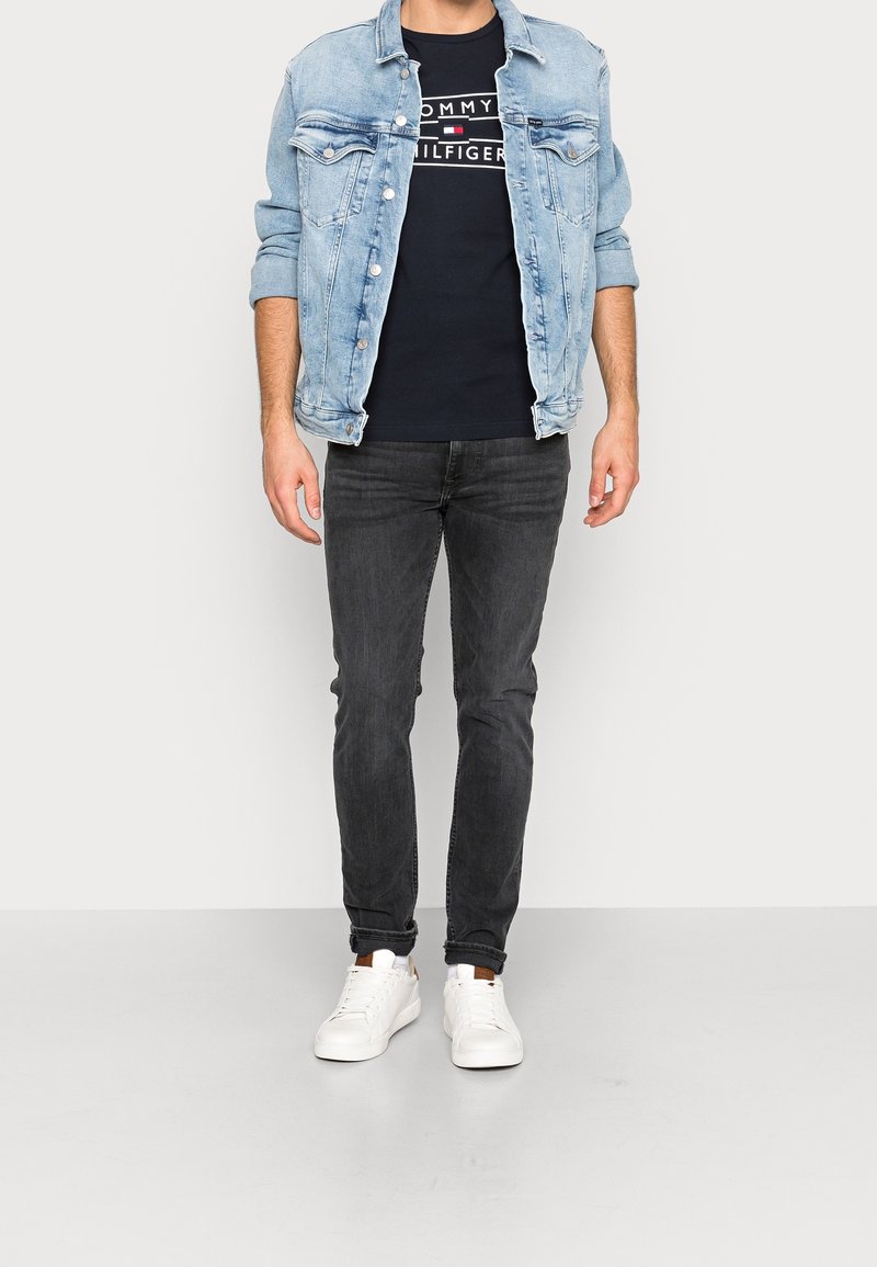 Lyseblå denimjakke over en navy Tommy Hilfiger t-shirt, parret med mørke jeans og hvide sneakers. Casual fit, minimalistisk design.