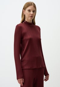 Bordeauxfarbener gerippter Rollkragenpullover mit langen Ärmeln, der dekorative goldene Knöpfe am Kragen und kontrastierende Seitennaht für visuelles Interesse aufweist.