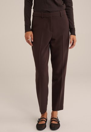 Hnědé kalhoty na míru se střihovým střihem, s plisováním a poutkem na opasek v pase, spárované s černými síťovanými botami na slip-on.