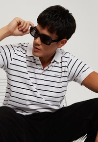 Biała koszulka polo w granatowe paski, z krótkimi rękawami i kołnierzykiem. Nosząca duże czarne okulary przeciwsłoneczne. Siedząca postawa na neutralnym tle.