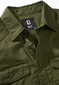 Camisa de algodón color verde oliva con botones, que presenta un cuello puntiagudo, dos bolsillos en el pecho y una etiqueta que indica la talla M.