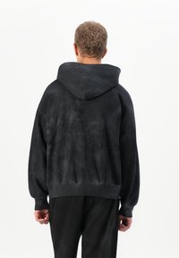 Person with short curly hair wearing a loose black hoodie and black pants, standing facing away against a plain white background.