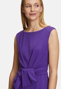 Lila ruha, puffos ujjakkal, redőzött kosárral és megkötős derékkal. Simább, textúrált anyag, amely testhezálló siluettet formáz.