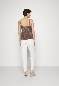 Camisola castanha com padrão e alças finas ajustáveis, emparelhada com calças brancas ajustadas e sandálias de salto de cor clara, vista de costas.