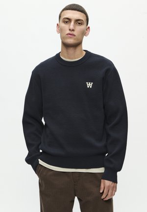 Navy knit sweater with ribbed crew neck and cuffs, featuring a small beige "W" logo on the chest, layered over a light beige shirt.