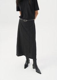 Fustă neagră, până la gleznă, cu croi drept, dintr-un material texturat, și cu o curea care are o cataramă rectangulară din argint. Purttată cu cizme negre cu toc.