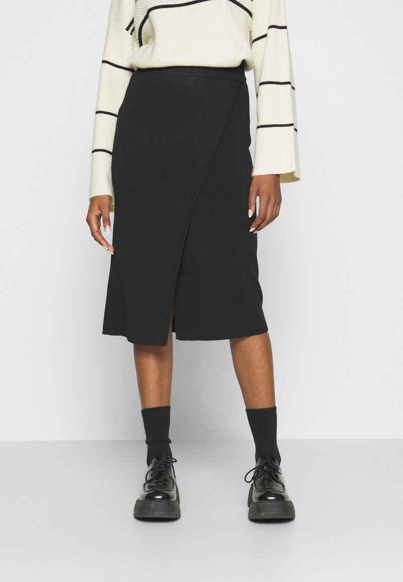 Femme portant une jupe portefeuille noire longueur genoux, des chaussettes noires, des chaussures noires à lacets et un pull blanc à rayures horizontales noires.