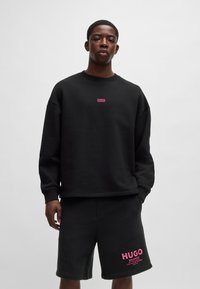 Felpa nera con finitura testurizzata, con un piccolo logo "HUGO" rosa. Abbinate a pantaloni corti neri coordinati, che mostrano anch'essi lo stesso logo in rosa.