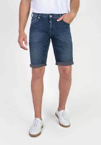 Pantalones cortos de mezclilla, azul oscuro, con un ajuste relajado y dobladillos enrollados. Equipados con bolsillos delanteros y traseros. Combinados con zapatillas blancas.