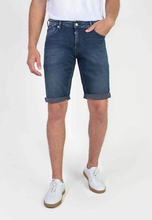 Pantalones cortos de mezclilla, azul oscuro, con un ajuste relajado y dobladillos enrollados. Equipados con bolsillos delanteros y traseros. Combinados con zapatillas blancas.