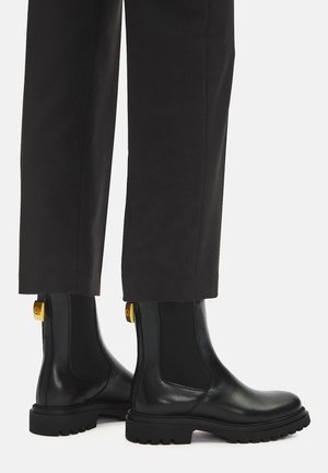 Black leather ankle boots with chunky rubber soles and elastic side panels, worn with black cropped pants against a white background.