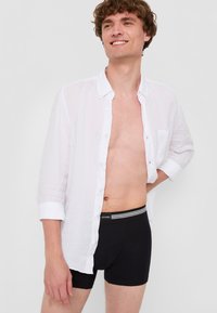 Vit linne skjorta med uppkavlade ärmar över svarta bomullsnattbyxor med randig midja. Ämnet ler, med en neutral bakgrund.