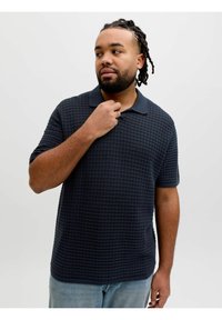 Homme avec cheveux tressés et barbe portant un polo à manches courtes texturé foncé et un jean bleu clair, tenant un collier et regardant sur le côté.