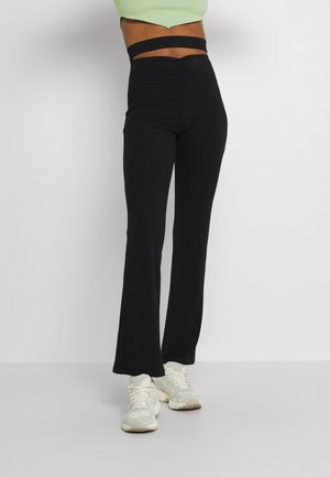 Pantalons évasés noirs à côtes avec une taille haute, présentant un design découpé à la taille. Portés avec des baskets de sport de couleur claire.