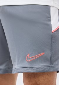 Γκρι αθλητικό σορτς με ροζ λογότυπο Nike swoosh, φορεμένα από άτομο, που δείχνουν υφασμάτινη πλέξη και μέρος λευκού αθλητικού μπλουζακίου.