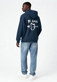 Námornícka mikina s bielou grafikou "Mi Amor" a dizajnom šípky na chrbte, skombinovaná so svetlomodrými džínsami a bielymi teniskami.