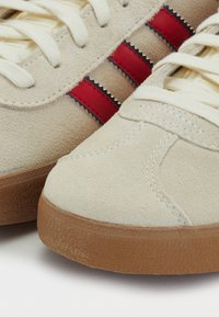 Sapatilhas de camurça bege com riscas vermelhas e brancas, apresentando uma sola de borracha texturizada e um design de biqueira redonda. Os atacadores são de cor creme.