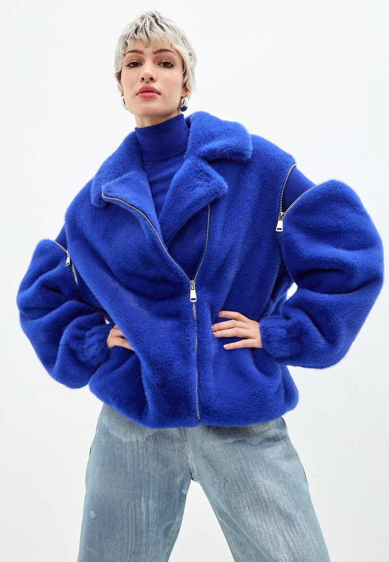 Giacca di pelliccia sintetica blu con colletto ampio, chiusura con zip e maniche oversize. Indossata sopra un dolcevita blu e abbinata a jeans chiari.