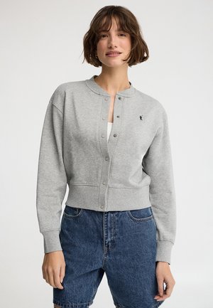 Mujer con cabello corto y castaño, vistiendo una sudadera gris claro abotonada y pantalones cortos de mezclilla, de pie frente a un fondo claro liso.