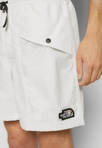 Short blanc avec poche à rabat frontale et bouton-pression noir, cordon noir, écusson logo "The North Face" près de l’ourlet, porté par une personne.
