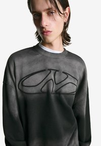 Jongere met halflang haar draagt een donkergrijze sweatshirt met een groot verheven "CK"-logo aan de voorkant, staande tegen een witte achtergrond.