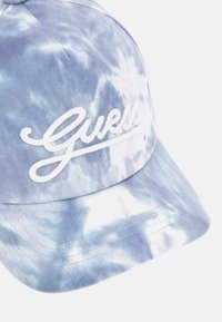 Gorra de tela con diseño de teñido azul y blanco, que presenta un logotipo bordado en blanco que dice "Guess" en la parte frontal, con visera curvada y detalles de costura.